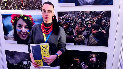 Dr. Anna Fomenko, mit roter Halskette und einem Programmheft der Cafe Kyiv Veranstaltung steht vor einer Bilderwand, die Eindrücke aus der neuen Lebensrealität ukrainischer Staatsbürger widerspiegeln. Zwischen Hoffnung und Leid.