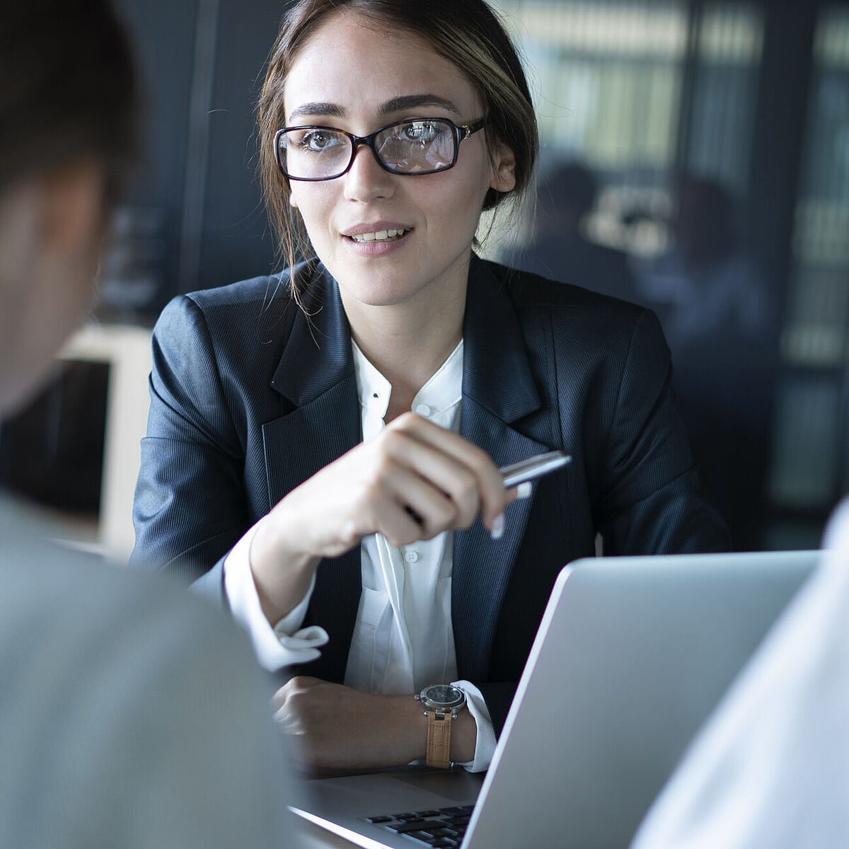 Eine Frau in Business Outfit und mit Brille sitzt vor zwei Personen und berät