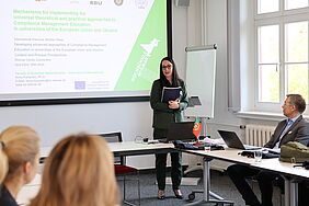 Die Projektkoordinatorin Frau Dr. Fomenko begrüßt ebenfalls die Teilnehmer, während hinter Ihr an der Leinwand die PowerPoint-Titelfolie für die Konferenz zu sehen ist.