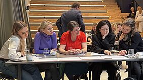 five women sitting at a table and looking at documents in front of them