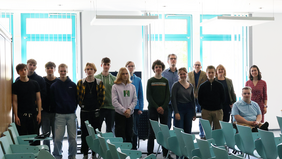 Gruppenfoto von rund 17 Personen, darunter Schüler:innen, Lehrkräfte und Hochschulangehörige, in einem Seminarraum mit türkisfarbenen Stühlen. Die Personen stehen und sitzen in zwei Reihen, vor hellen Fenstern.