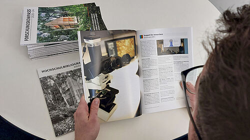 You look over the shoulder of a person holding an unfolded brochure in their hands. On the table are more copies and an insert that is slightly smaller than the brochure printed in A4 format.