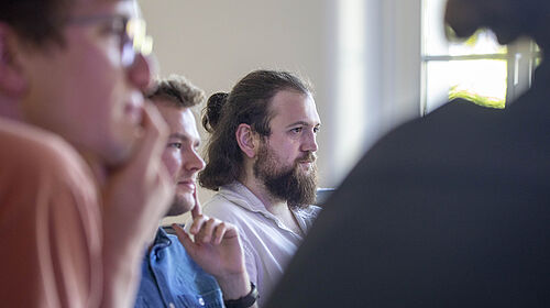 Drei junge Promovierende sitzen nebeneinander in einem Seminarraum und hören aufmerksam zu. Der Mann in der Mitte trägt einen Vollbart und hat lange Haare, die zu einem Knoten gebunden sind. Die anderen beiden Männer tragen Brillen und haben den Blick nach vorne gerichtet.