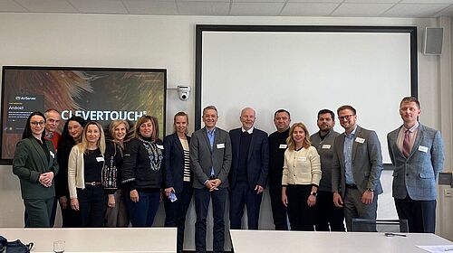 13 Personen stehen nebeneinander. Im Hintergrund ist eine große Leinwand und links davon ein großer Smart-Touch Bildschirm zu sehen.