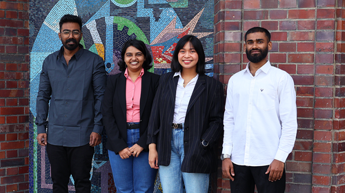 Four people stand next to each other in front of a brick wall with a colourful mosaic design. They are all looking at the camera and smiling. The people are wearing business or casual clothing.