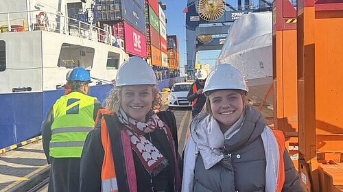 Auf dem Bild sind mittig zwei Frauen. Sie tragen Warnwesten und Sicherheitshelme auf ihrem Kopf. Im Hintergrund ist ein Containerschiff zu sehen. Die Frauen lächeln.